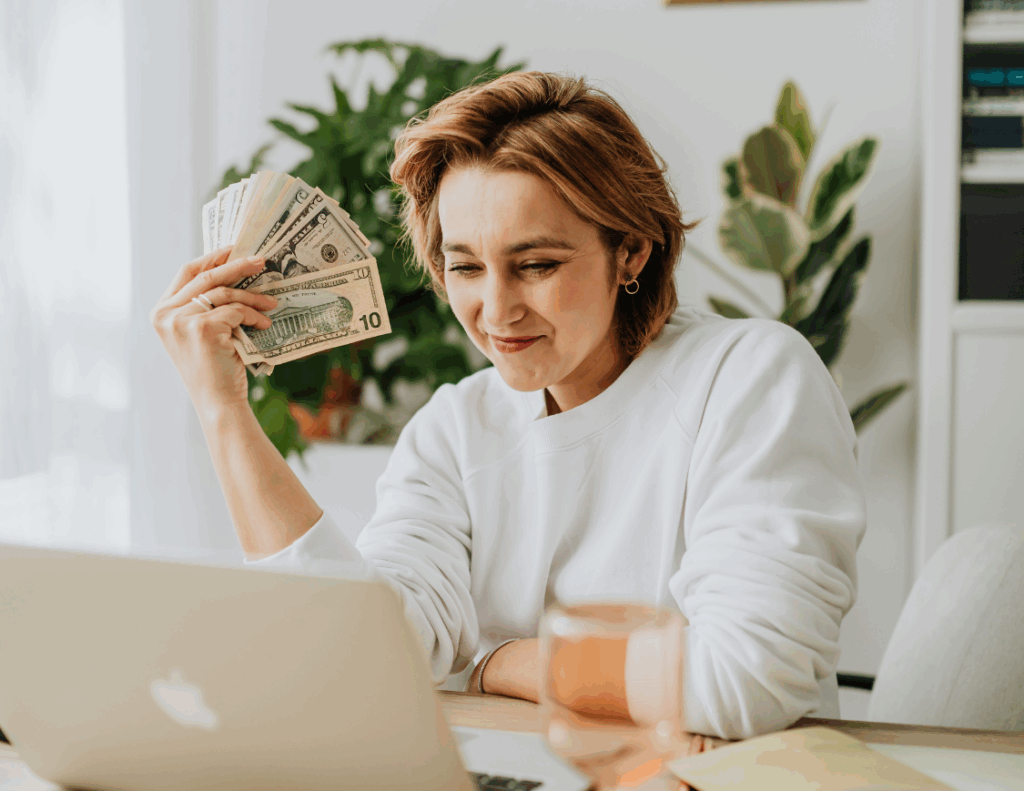 Femme souriante devant son ordinateur tenant de l’argent en main après réception d’une facture réglée.