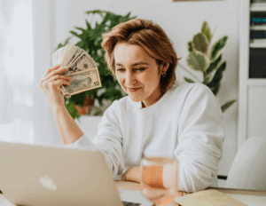 Femme souriante devant son ordinateur tenant de l’argent en main après réception d’une facture réglée.